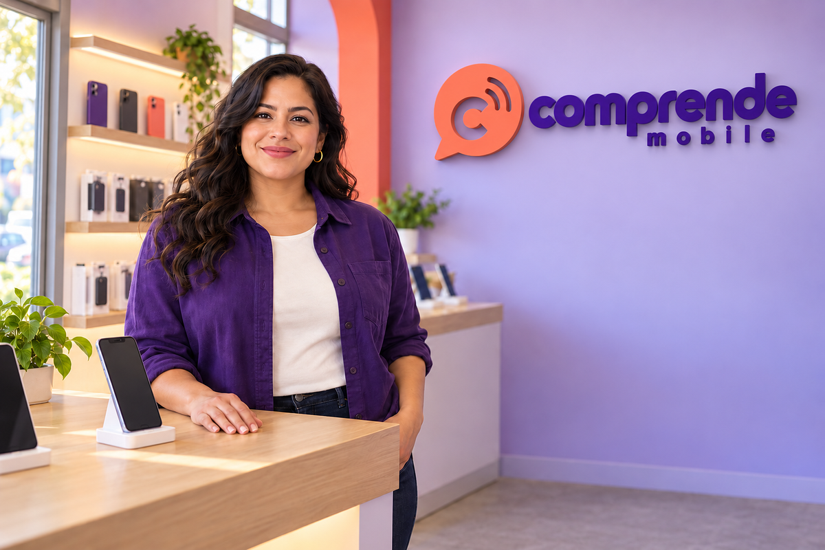 A store owner smiling behind the counter of a bright phone shop with a Comprende Mobile sign on the wall.